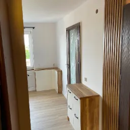 Nowoczesny przedpokój z drewnianymi lamelami na ścianie, białą komodą z drewnianym blatem i czarnymi uchwytami oraz drzwiami wewnętrznymi z dekoracyjną szybą.