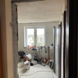 BUILDMEB Paweł Włodarczyk - Wnętrze w trakcie demontażu ścianki działowej, widoczne gruzowisko, okno z widokiem na zabudowę jednorodzinną i drzwi wewnętrzne z szybą w folii ochronnej.