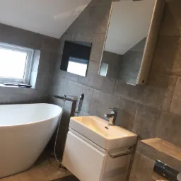 Nowoczesna łazienka z wolnostojącą wanną, wiszącą szafką z umywalką, lustrem z oświetleniem i szarą płytką imitującą kamień na ścianach i podłodze.