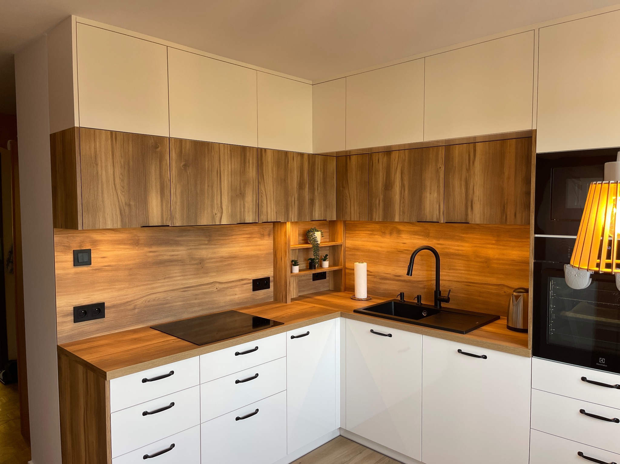 Nowoczesna kuchnia z białymi szafkami dolnymi i drewnianymi górnymi, czarny zlew i bateria, drewniany panel ścienny z półkami na rośliny, płyta indukcyjna, piekarnik Electrolux i lampa...