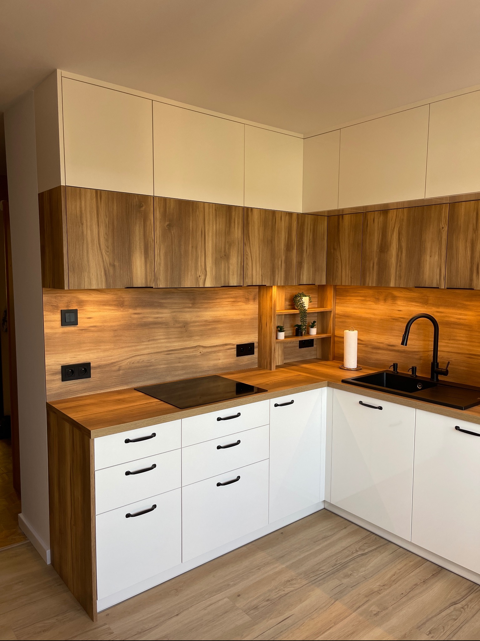 Nowoczesna kuchnia z białymi szafkami dolnymi i drewnopodobnymi górnymi, czarny zlew i bateria, drewniany blat, podświetlenie LED pod szafkami, minimalistyczny design, rośliny doniczkowe na półce.