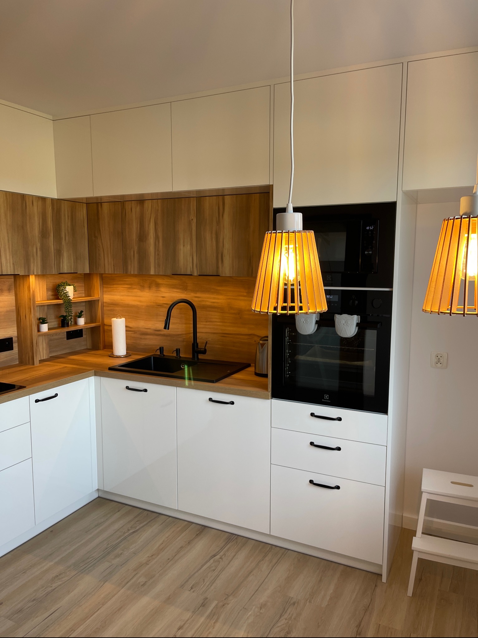 Nowoczesna kuchnia z białymi i drewnianymi frontami, czarnym zlewem i baterią, oświetlona lampami z drewnianymi abażurami, z piekarnikiem i mikrofalówką w zabudowie.