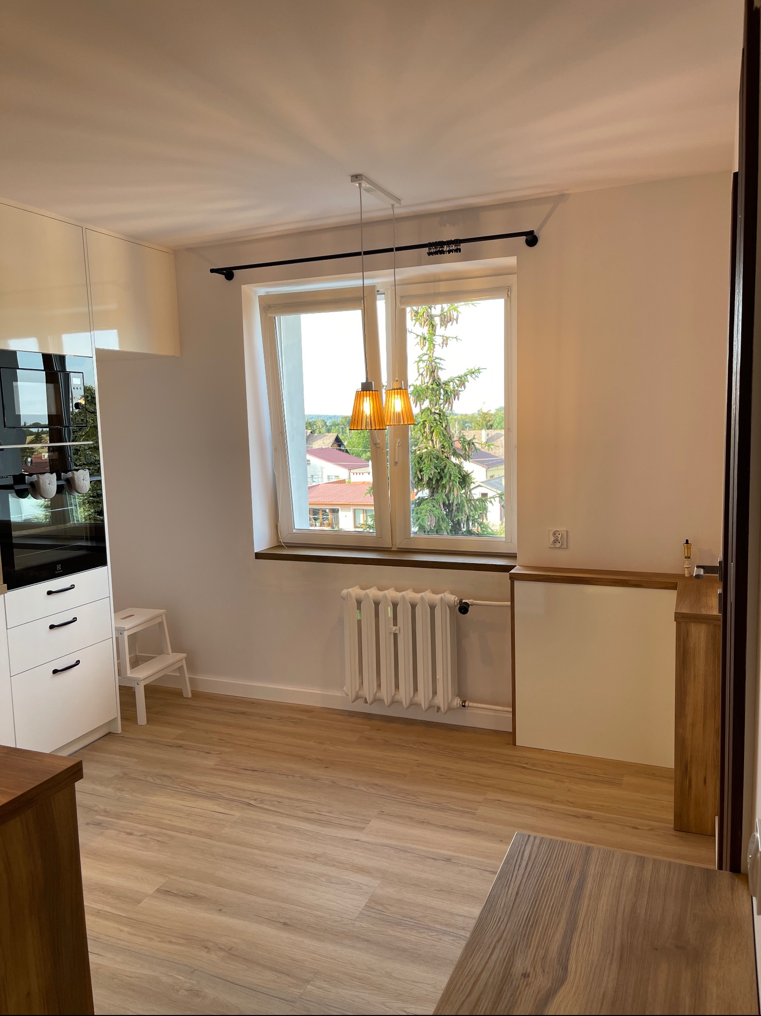 Jasna kuchnia z białymi szafkami, drewnianym blatem i podłogą, widok na drzewo za oknem i wisząca lampa z abażurami w kolorze miodowym.