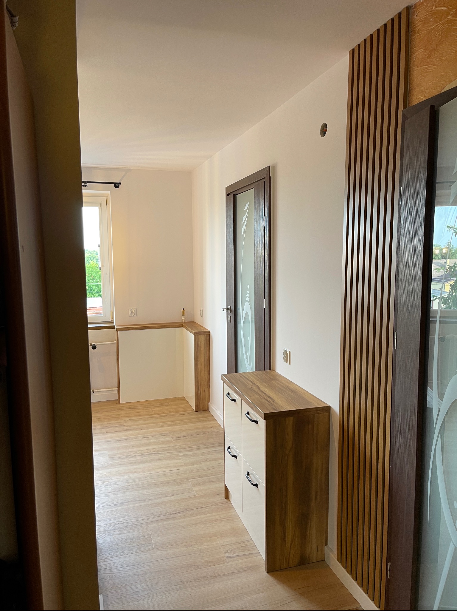 Nowoczesny przedpokój z drewnianą zabudową, białą komodą z drewnianym blatem, lamelami na ścianie i przeszklonymi drzwiami, widok z korytarza.