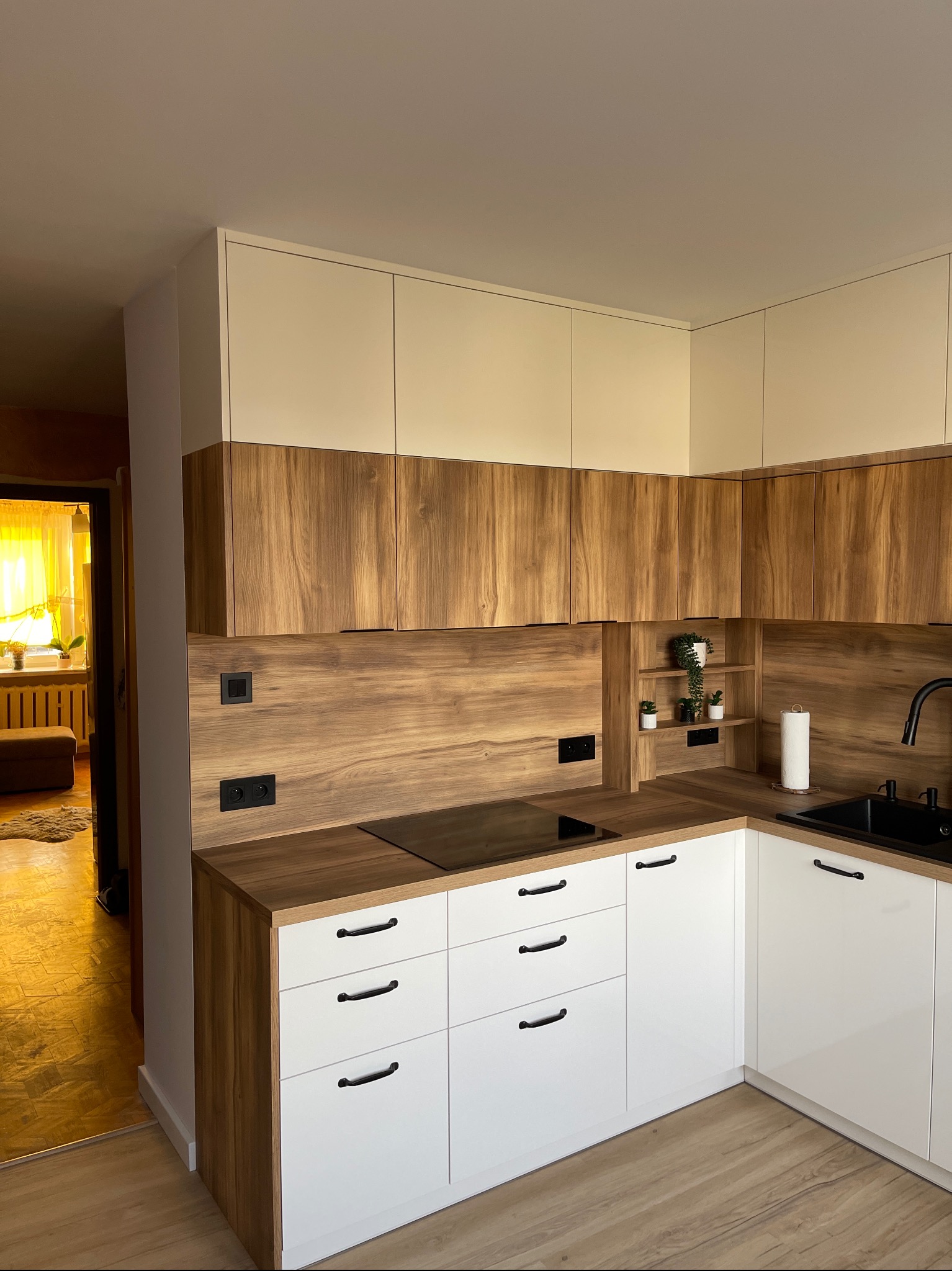 Nowoczesna kuchnia narożna z białymi szafkami dolnymi i drewnopodobnymi szafkami górnymi, czarnym blatem i armaturą, widok na sąsiedni pokój z żółtymi zasłonami.