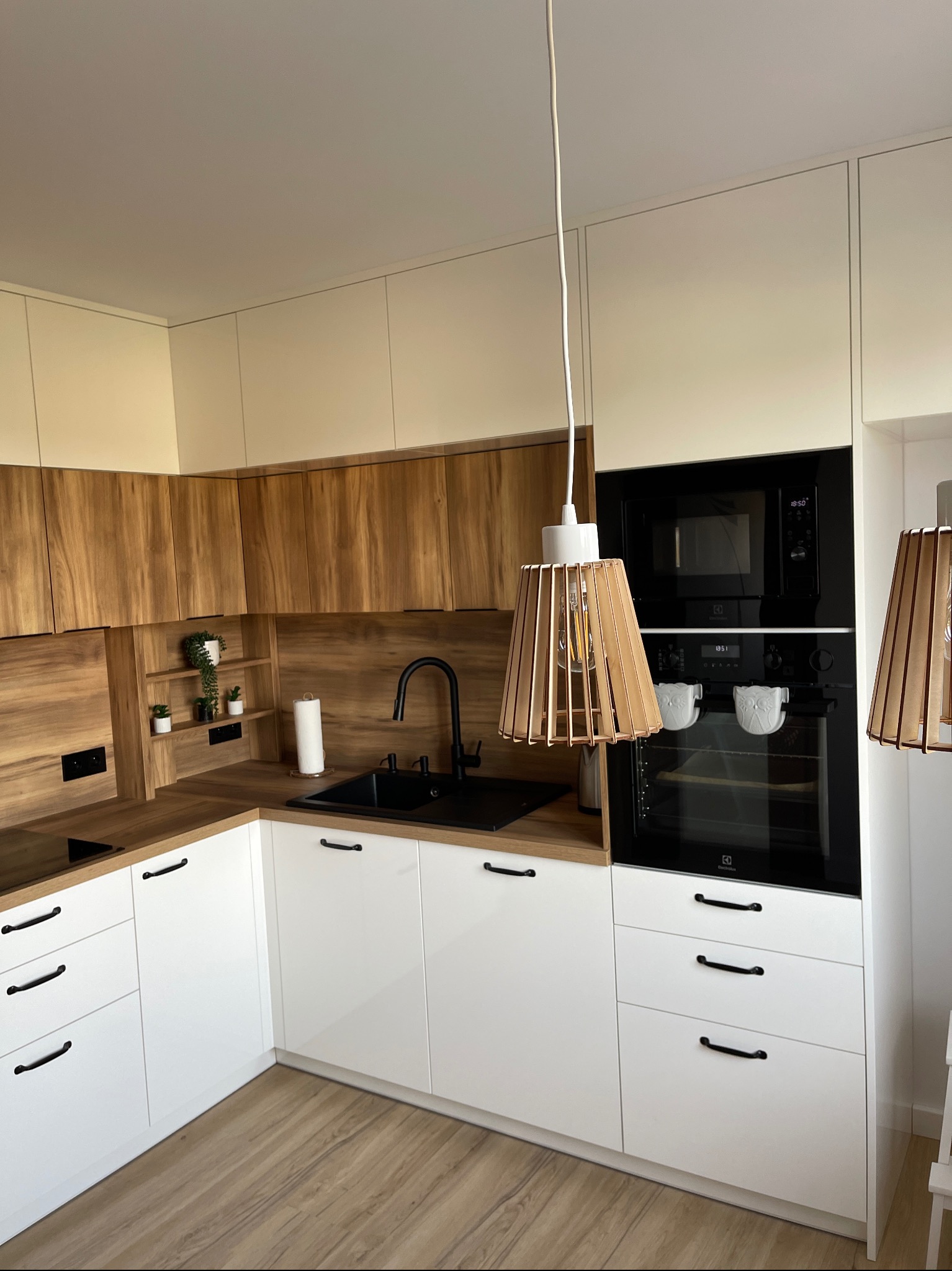Nowoczesna kuchnia z drewnianymi szafkami górnymi, białymi dolnymi, czarnym zlewem i baterią, oraz drewnianymi lampami wiszącymi. W zabudowie piekarnik i mikrofalówka.