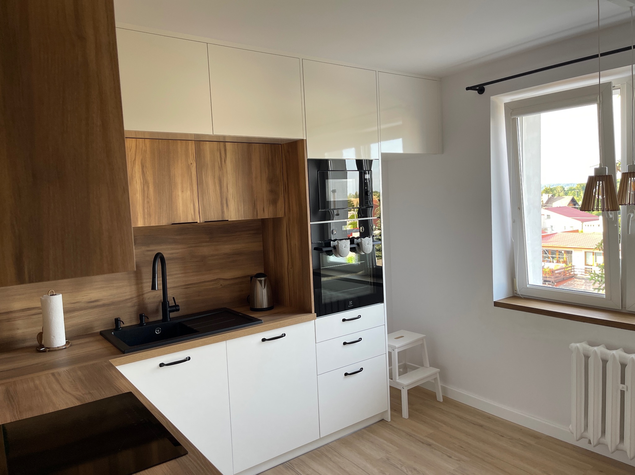 Nowoczesna kuchnia z białymi i drewnianymi frontami, czarną armaturą, piekarnikiem Electrolux i widokiem na osiedle przez okno.