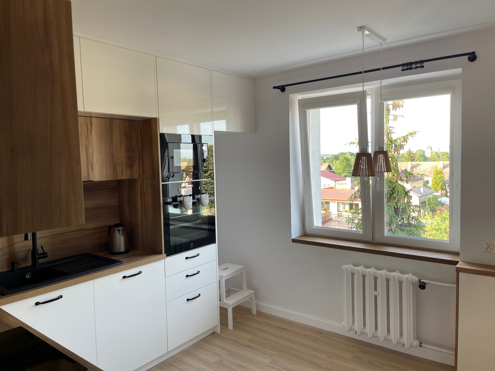 Jasna kuchnia z drewnianymi i białymi szafkami, czarnym zlewem i baterią, oraz oknem z widokiem na osiedle domów jednorodzinnych.