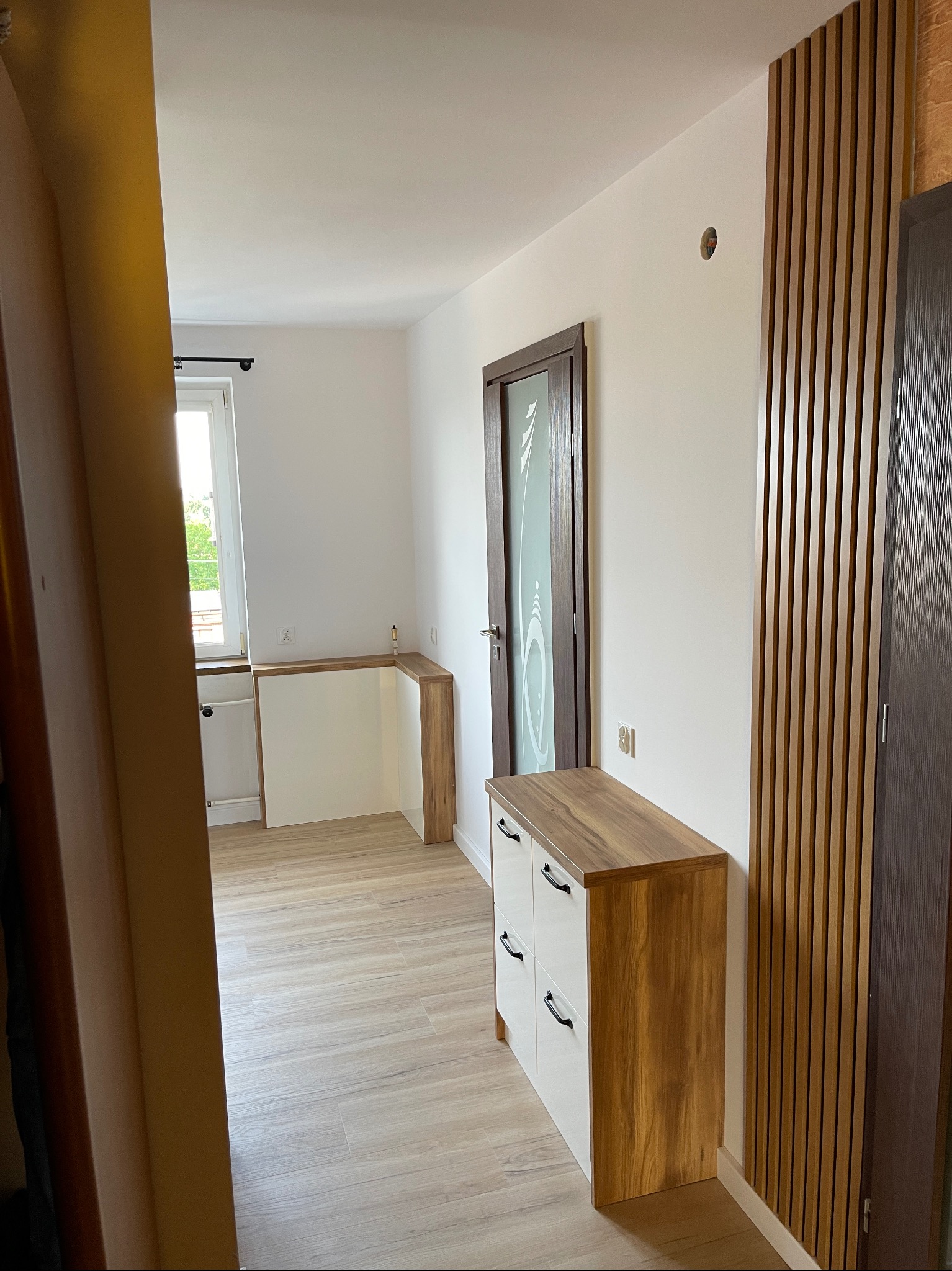 Nowoczesny przedpokój z drewnianymi lamelami na ścianie, białą komodą z drewnianym blatem i czarnymi uchwytami oraz drzwiami wewnętrznymi z dekoracyjną szybą.