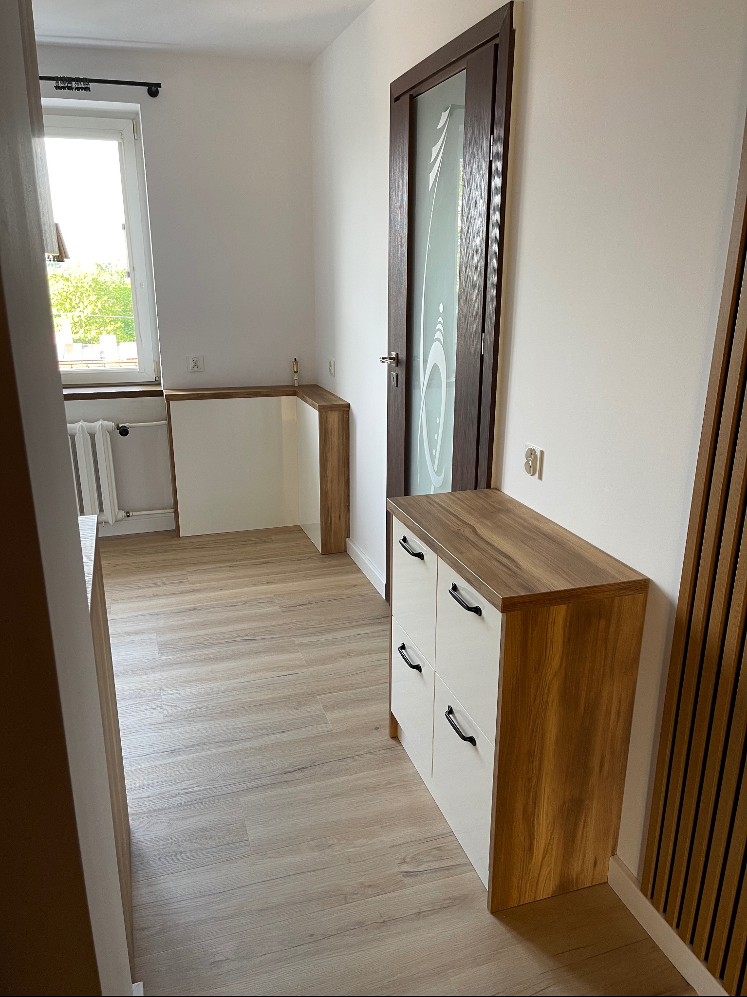 Nowoczesna szafka z białymi frontami i drewnianym blatem w przedpokoju, obok drzwi z matowym szkłem i dekoracyjnej ściany z drewnianych listew.