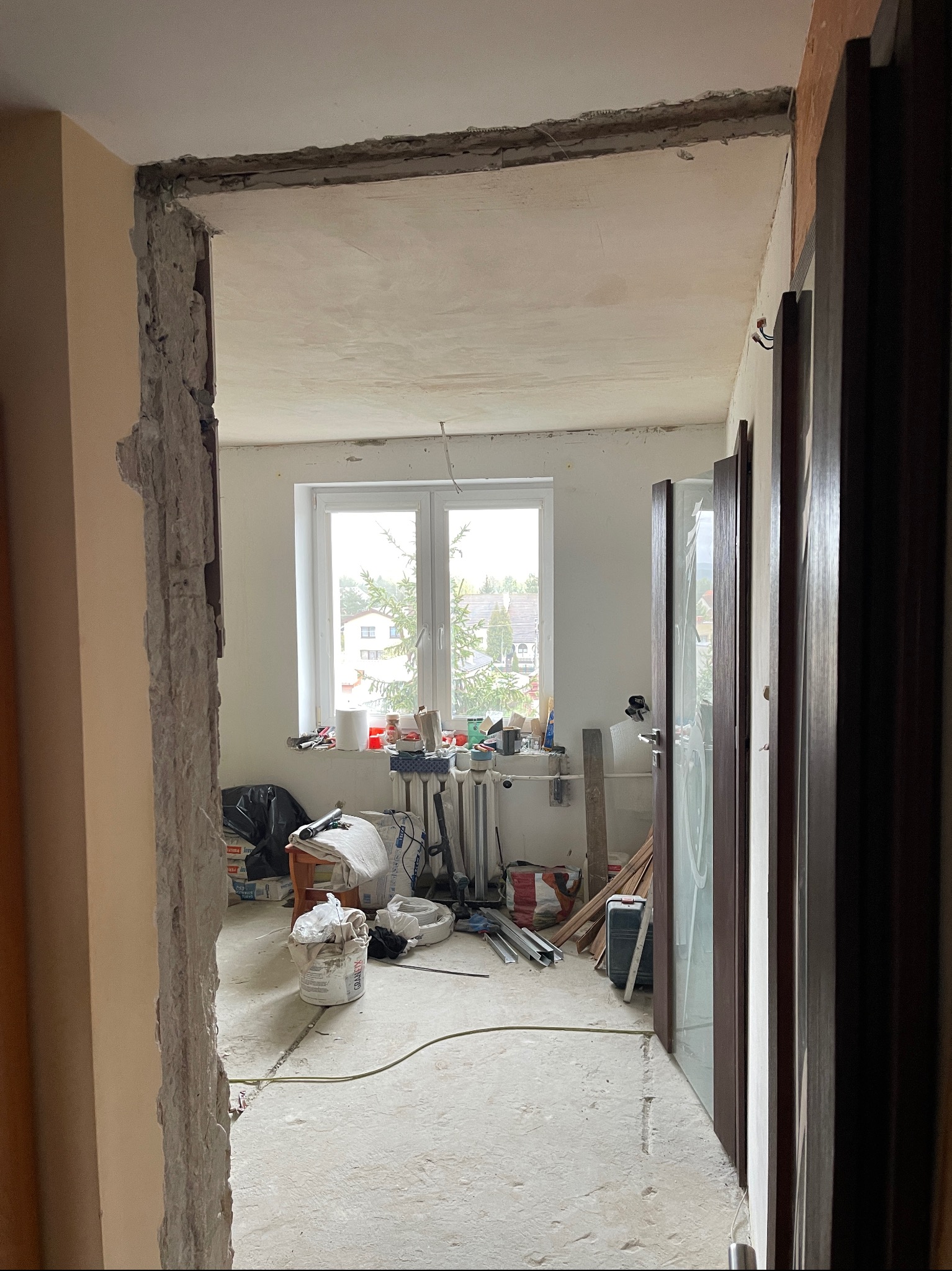 Wnętrze w trakcie demontażu ścianki działowej, widoczne gruzowisko, okno z widokiem na zabudowę jednorodzinną i drzwi wewnętrzne z szybą w folii ochronnej.