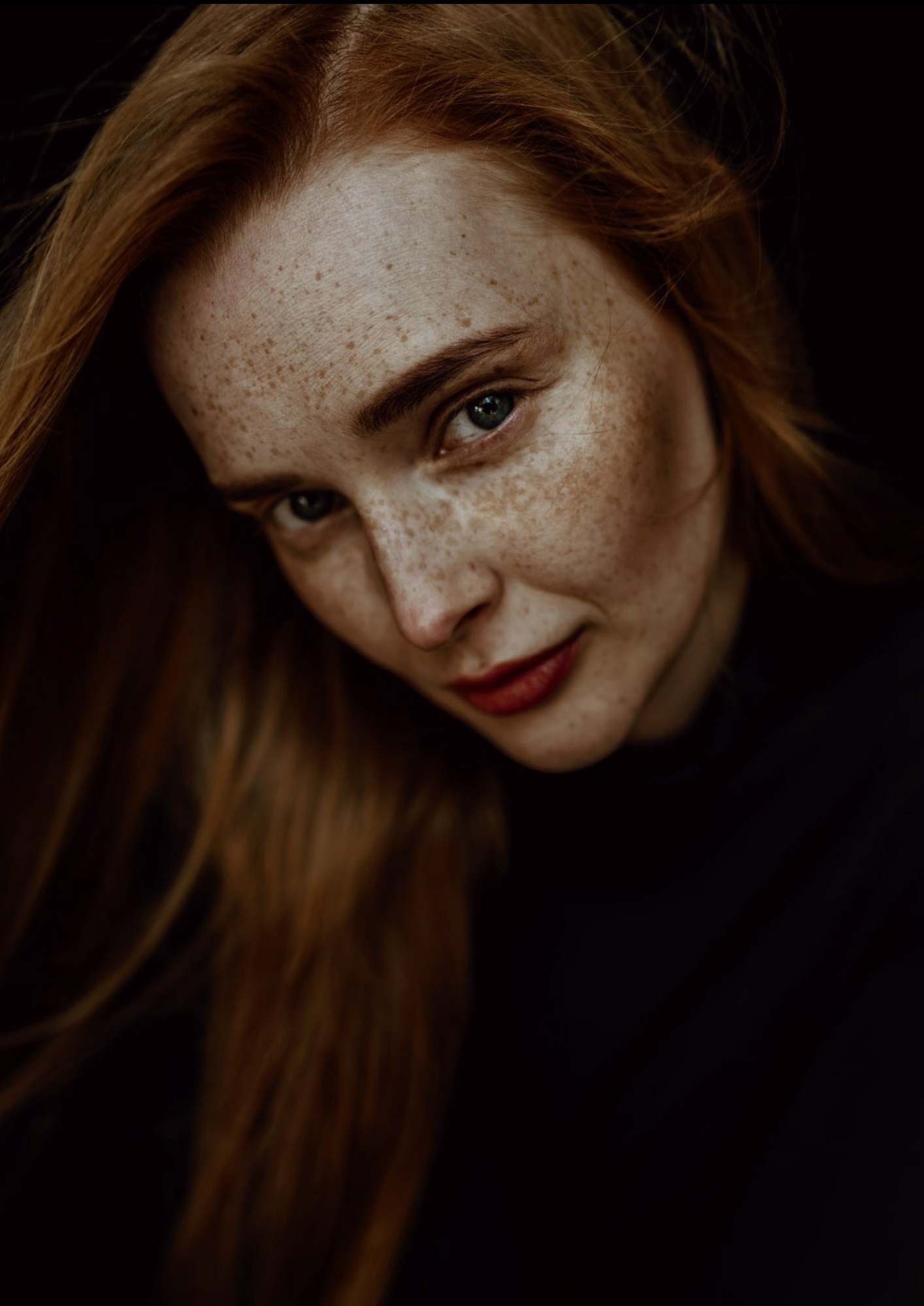 Portret kobiety z rudymi włosami i piegami, delikatny makijaż z czerwoną szminką, ciemne tło.