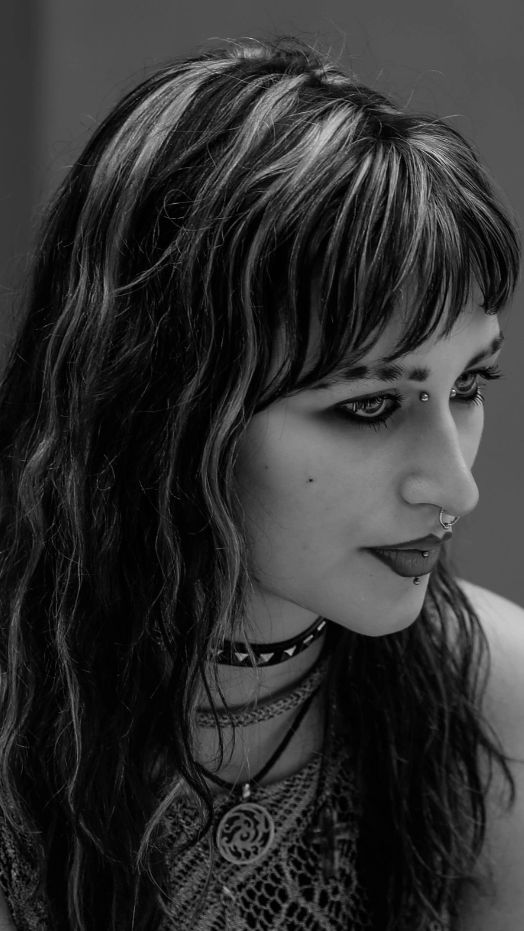 Czarno-biały portret kobiety z piercingiem na twarzy, ciemnym makijażem oczu i ust, oraz chokerem z ćwiekami i naszyjnikiem z okrągłym wisiorkiem.