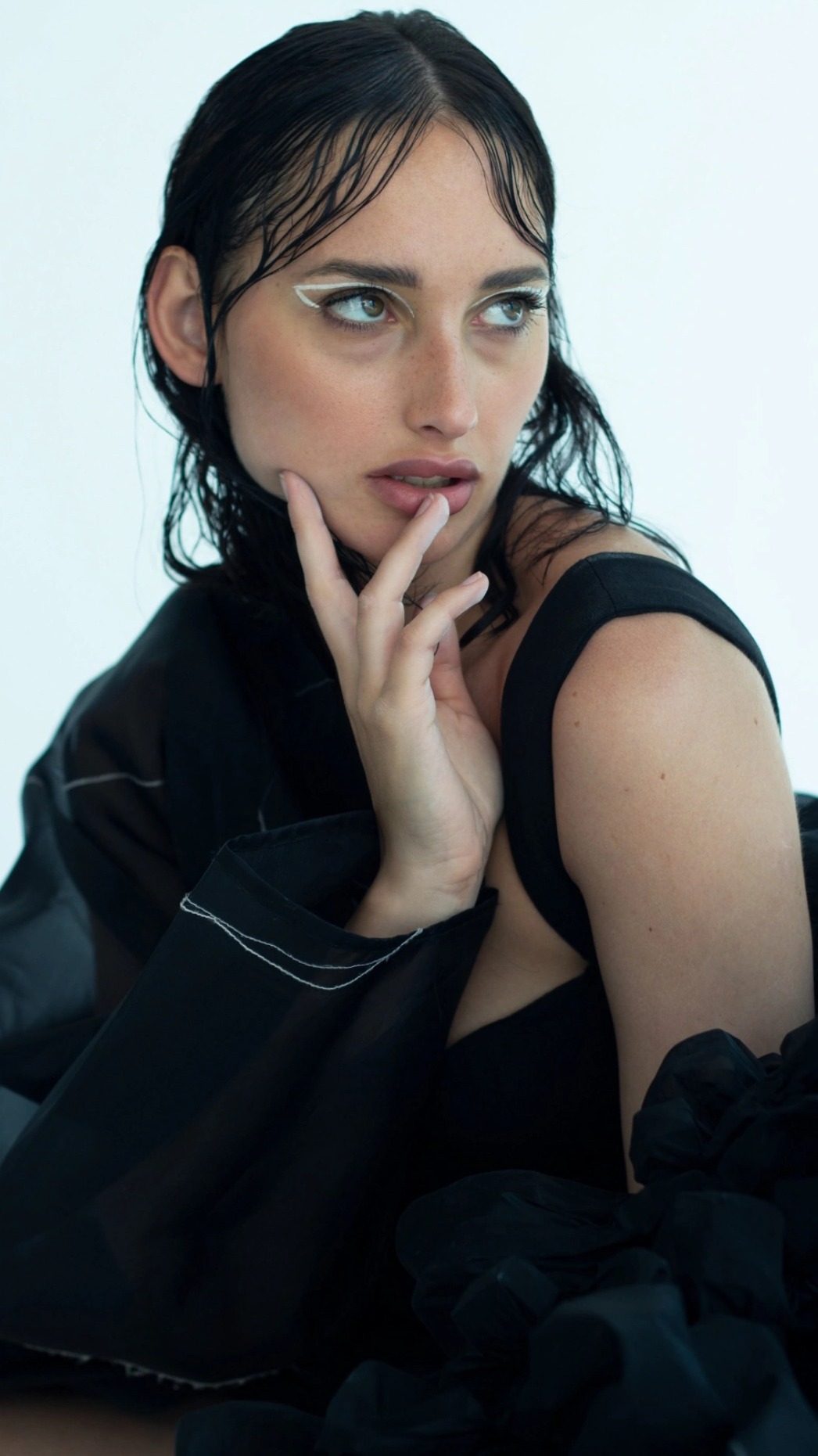 Portret kobiety z mokrymi włosami, w czarnej sukience z odkrytymi ramionami, z białym eyelinerem, delikatnie dotykającej ust palcami, zamyślony wyraz twarzy.