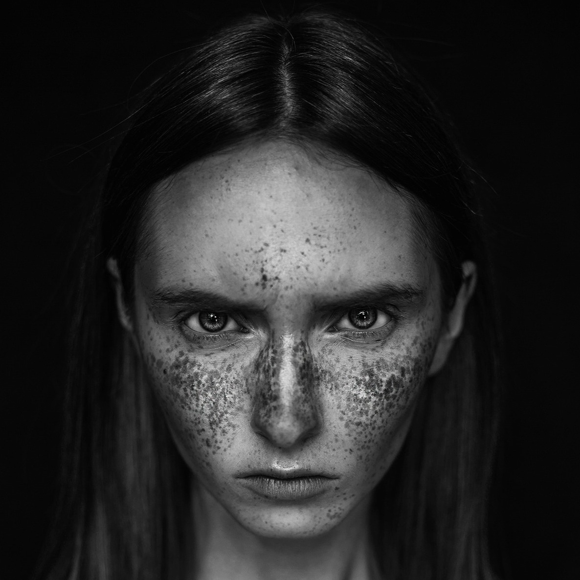 Czarno-biały portret kobiety z intensywnym spojrzeniem i wyraźnymi piegami na twarzy, oświetlonej z góry.