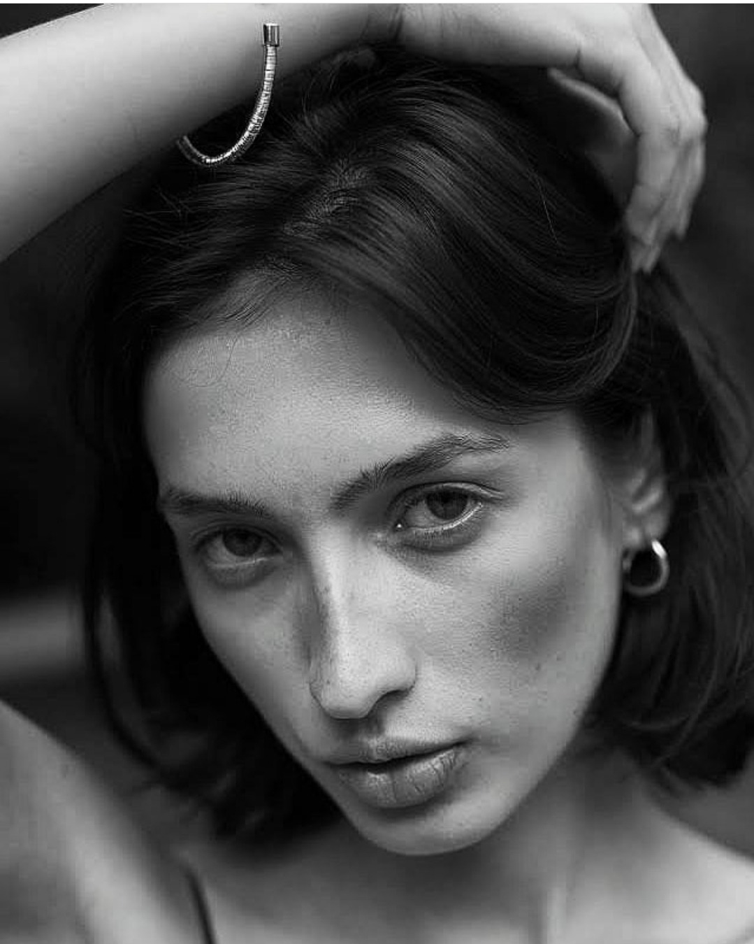 Portret kobiety w czerni i bieli, z bransoletką na ramieniu, skierowana w dół, z subtelnym makijażem i kolczykiem w uchu.