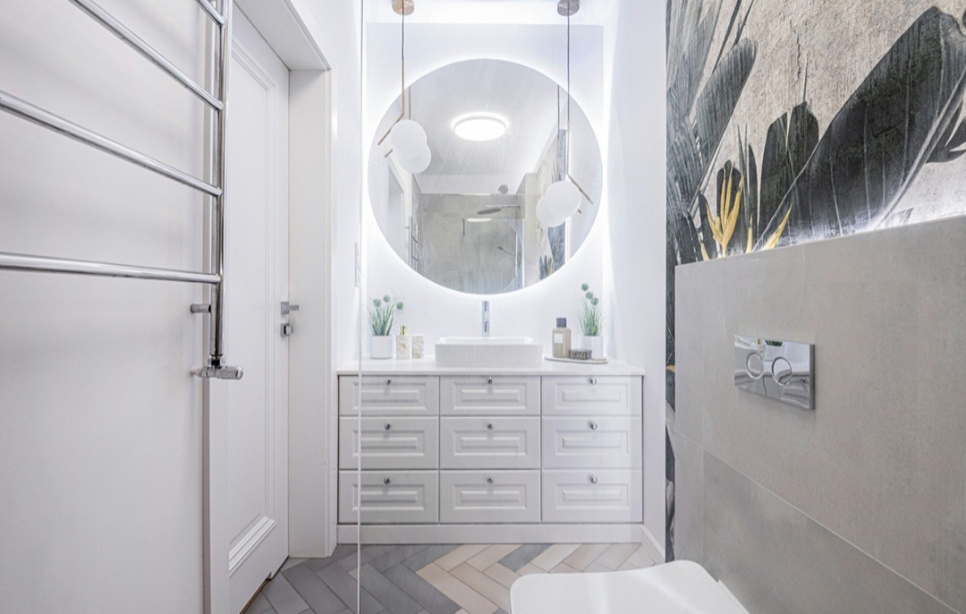 Jasna łazienka z okrągłym lustrem podświetlanym LED, białą szafką z szufladami, umywalką nablatową i grzejnikiem drabinkowym.