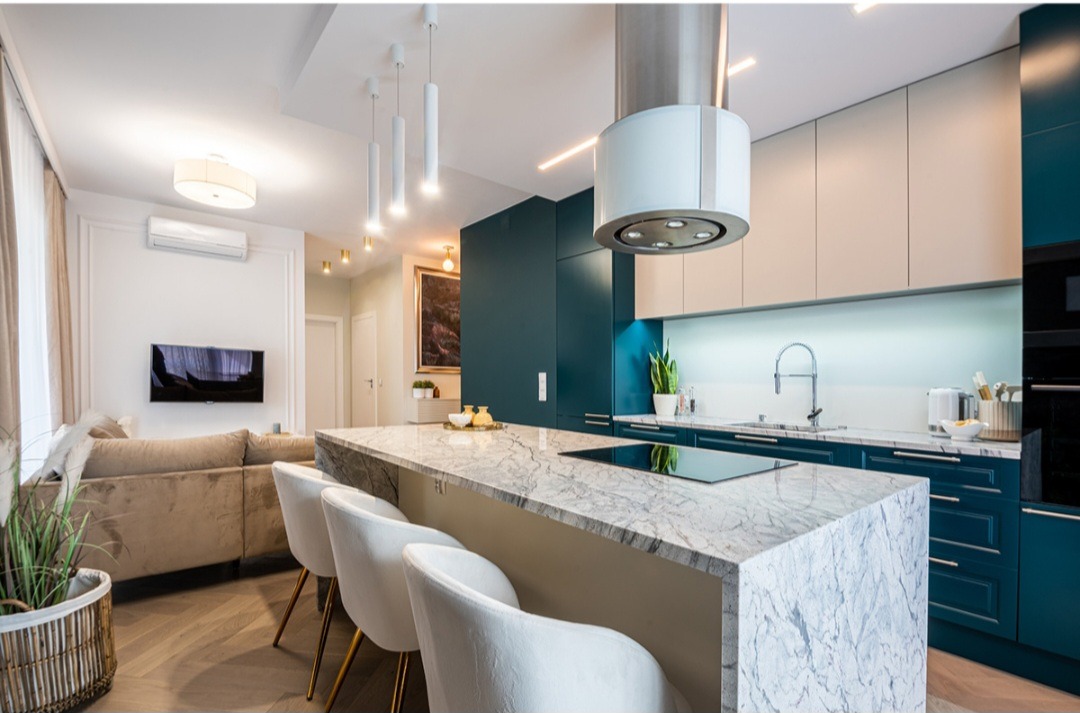 Nowoczesna kuchnia z wyspą o marmurowym blacie, białe krzesła barowe z złotymi nogami, ciemnozielone szafki, designerski okap i oświetlenie.