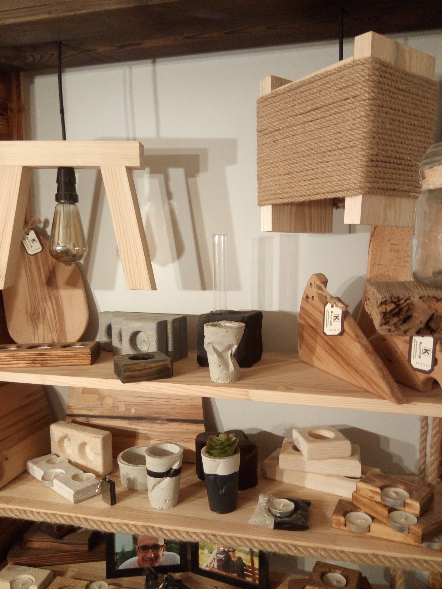 Półka z wyrobami z drewna: lampa na drewnianym trójnogu, drewniane deski do krojenia, doniczki z betonu architektonicznego, drewniane świeczniki i inne dekoracje.