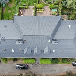 Widok z drona na duży dom z nowym, grafitowym dachem z dachówki ceramicznej, z licznymi oknami dachowymi i trzema lukarnami, otoczony zadbanym ogrodem i trzema samochodami.