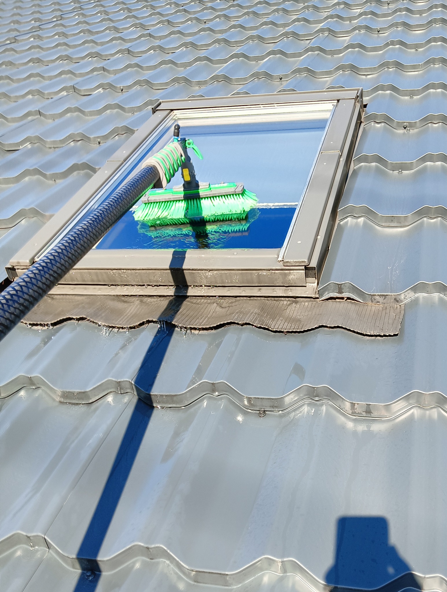 Czyszczenie okna dachowego na metalowej dachówce za pomocą szczotki na teleskopowym kiju z dopływem wody. Widoczne krople wody na dachu i oknie.