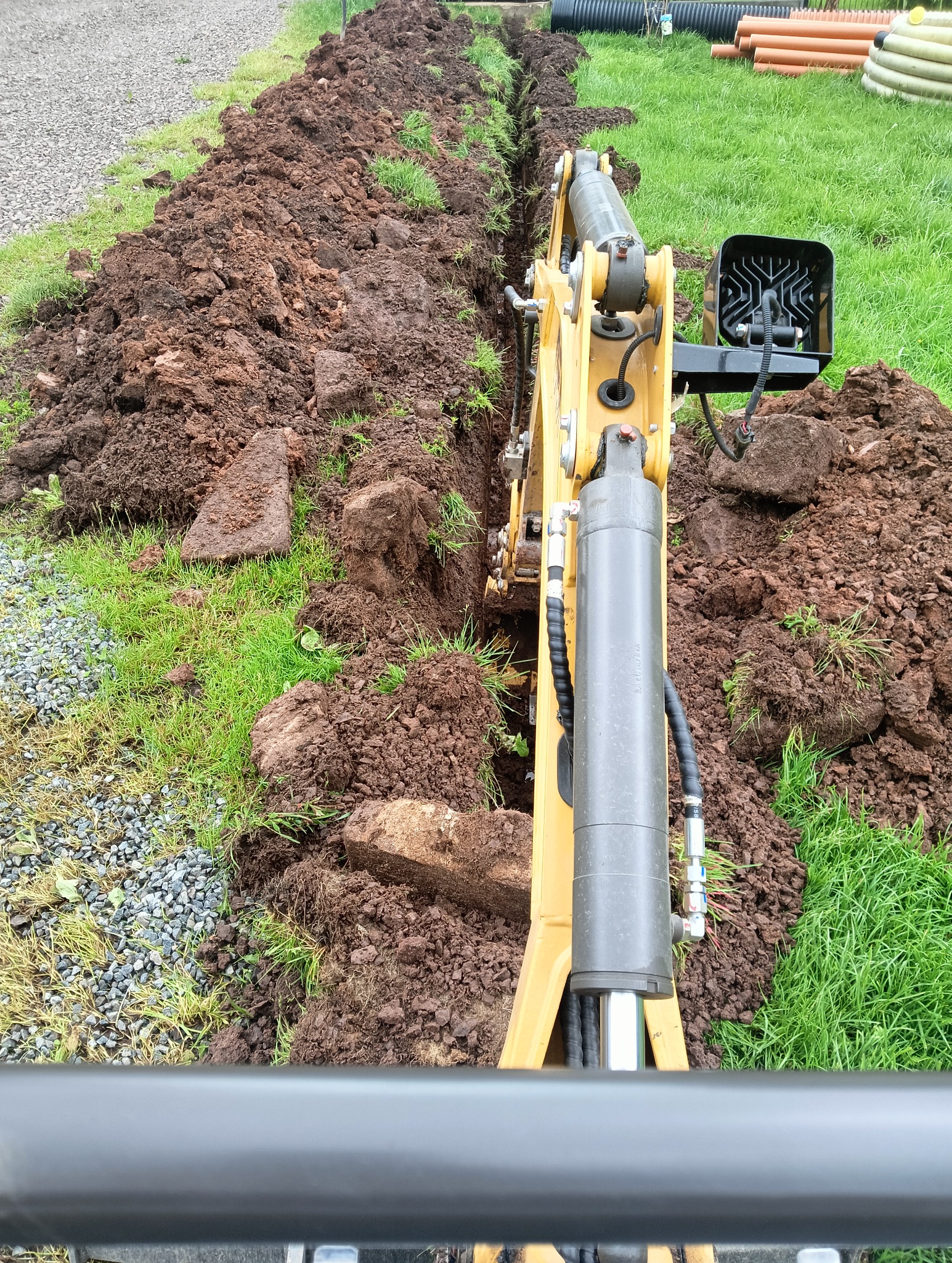 Żółta minikoparka w trakcie wykonywania wykopu pod instalacje w ziemi. Widoczna świeżo wykopana bruzda w ziemi i rury w tle. Prace ziemne.