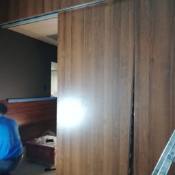 Golden Hands - Montaż przesuwanych drzwi z drewnopodobnej okleiny w biurze, widoczny pracownik w niebieskiej koszulce i aluminiowa drabina oparta o ścianę.