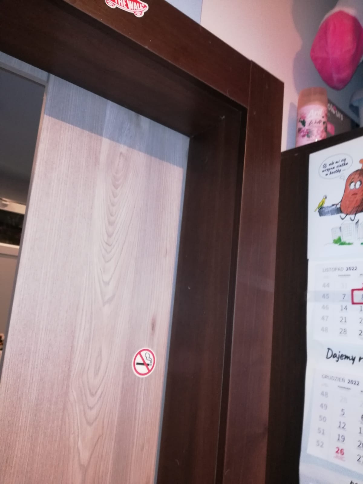 Drewniana ościeżnica z naklejką 'zakaz palenia' na jasnych drzwiach, widoczny fragment kalendarza na ścianie i naklejka Vans na ościeżnicy.