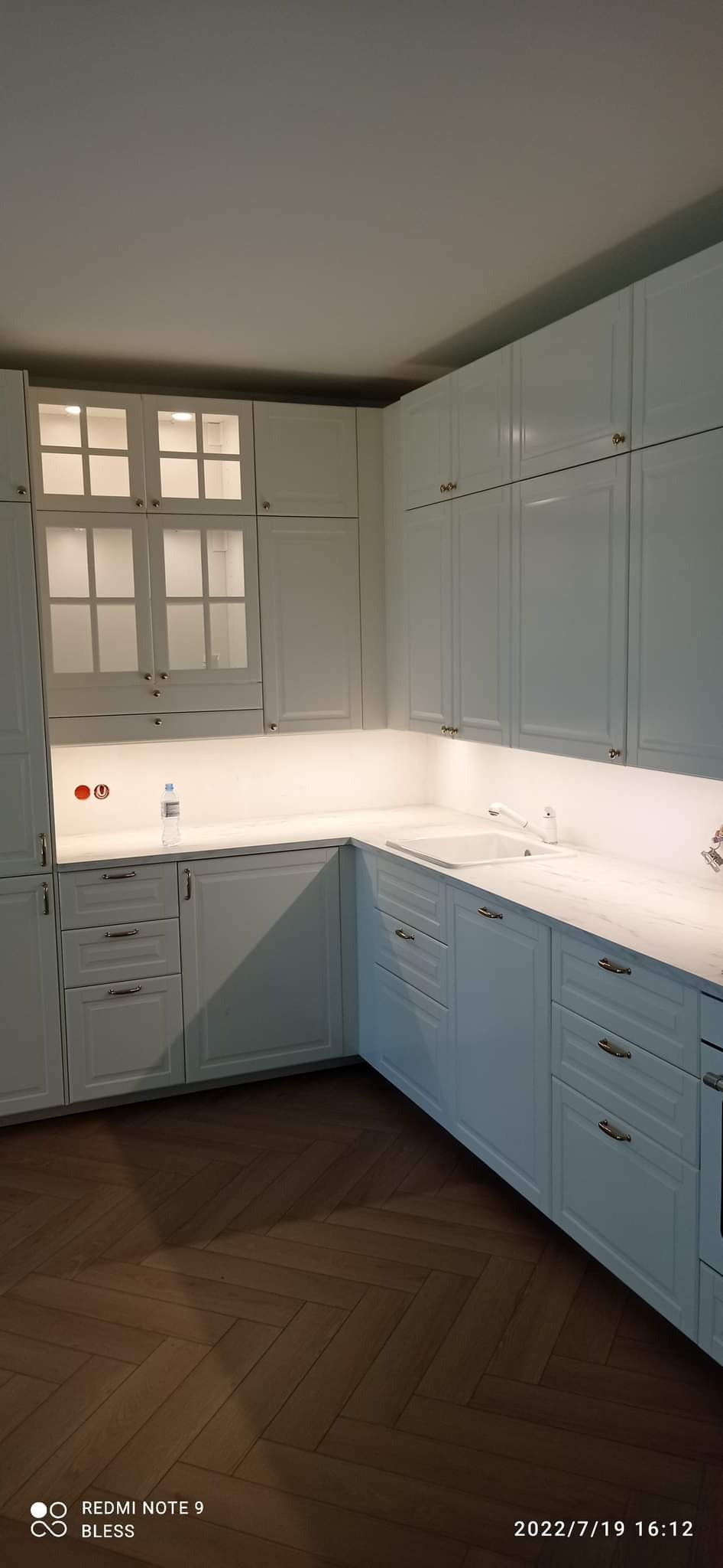 Kuchnia w stylu klasycznym z białymi szafkami, marmurowym blatem i drewnianą podłogą w jodełkę; widoczne oświetlenie podszafkowe i szklane witryny w górnych szafkach.