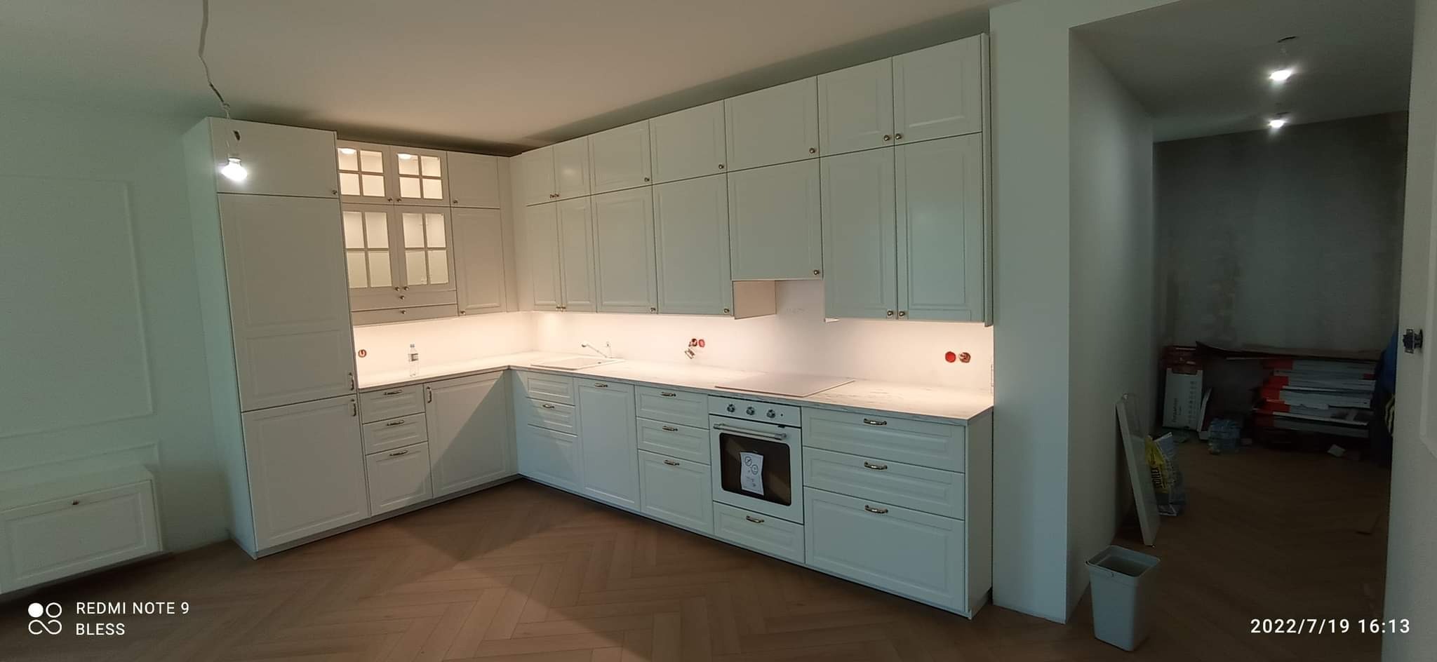 Jasna, klasyczna kuchnia z białymi szafkami, marmurowym blatem i oświetleniem podszafkowym, parkiet w jodełkę na podłodze, widok na przyległe pomieszczenie w trakcie prac wykończeniowych.