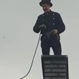 Kominiarz w tradycyjnym stroju i cylindrze, uśmiechnięty, stoi na dachu obok komina z cegły, trzymając narzędzie do czyszczenia komina.