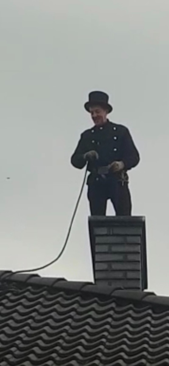 Kominiarz w tradycyjnym stroju i cylindrze, uśmiechnięty, stoi na dachu obok komina z cegły, trzymając narzędzie do czyszczenia komina.