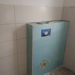 Niedokończona zabudowa stelaża podtynkowego WC z płyt karton-gips, widoczne przyłącza wodne, obok ściana wyłożona beżowymi płytkami, na górze poziomnica i nóż.