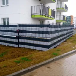 Nowoczesne ogrodzenie z panelami w kolorze antracytu i szarości, zamontowane przed nowym budynkiem mieszkalnym z balkonami w kolorze limonki, widoczne z perspektywy chodnika.