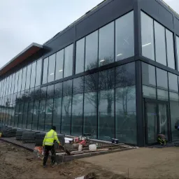 Montaż kostki brukowej przed nowoczesnym budynkiem z przeszkloną fasadą i czarnymi ramami okiennymi; pracownicy w odblaskowych kamizelkach układają kostkę, widoczne narzędzia i materiały budowlane.