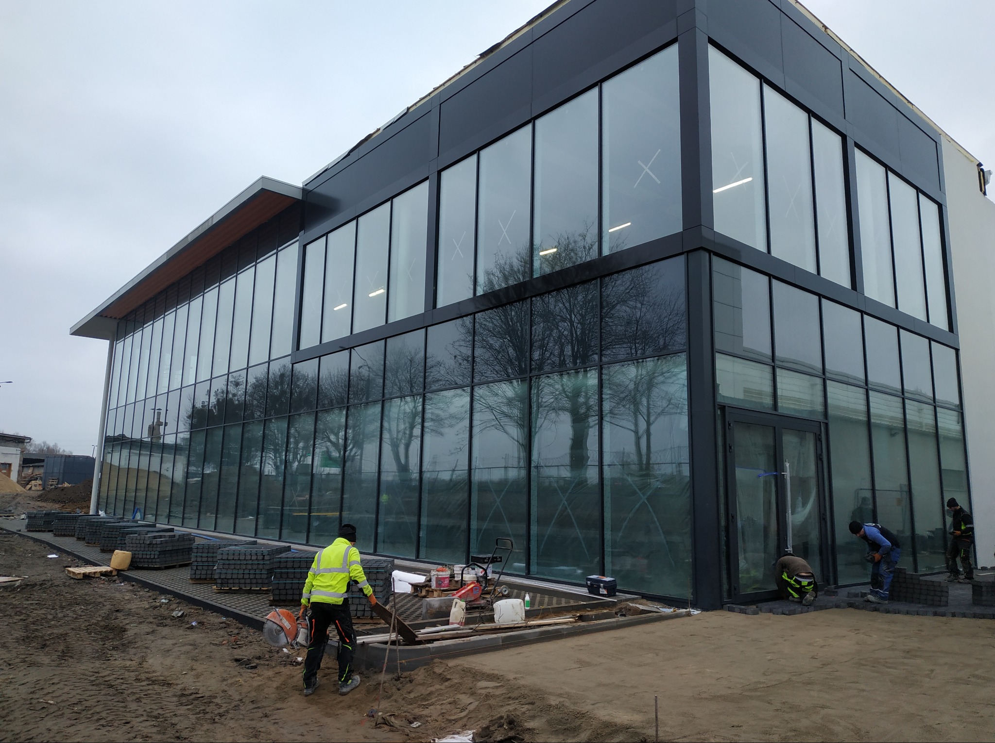 Montaż kostki brukowej przed nowoczesnym budynkiem z przeszkloną fasadą i czarnymi ramami okiennymi; pracownicy w odblaskowych kamizelkach układają kostkę, widoczne narzędzia i materiały budowlane.