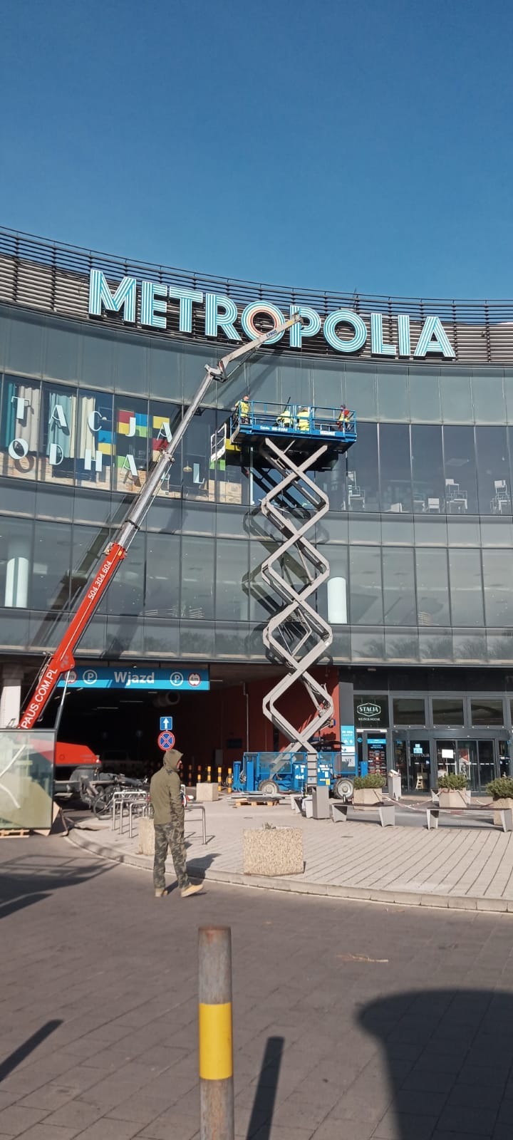 Pracownicy na podnośniku koszowym montują podświetlany napis METROPOLIA na fasadzie budynku, widoczny fragment wjazdu do parkingu i przechodząca osoba.