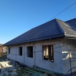 Energo-Dom - Dom w trakcie budowy: ściany z białych bloczków, ciemnoszary dach z rynną, widoczne otwory okienne, gruz i materiały budowlane na pierwszym planie, błękitne niebo w tle.