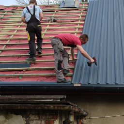 Energo-Dom - Dwóch dekarzy montuje blachodachówkę na drewnianej konstrukcji dachu; jeden przykręca wkrętarką arkusz blachy, drugi stoi w tle na łatach pokrytych czerwoną folią.