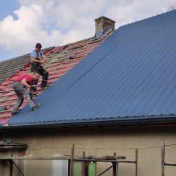 Energo-Dom - Dwóch dekarzy montuje blachodachówkę w kolorze grafitowym na połaci dachu, w tle widoczny komin z cegły i częściowo zdjęte stare pokrycie.
