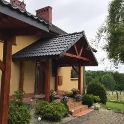 Żółty dom z ciemnym, spadzistym dachem i drewnianym zadaszeniem nad wejściem, ozdobiony kwiatami w donicach i zadbaną zielenią.