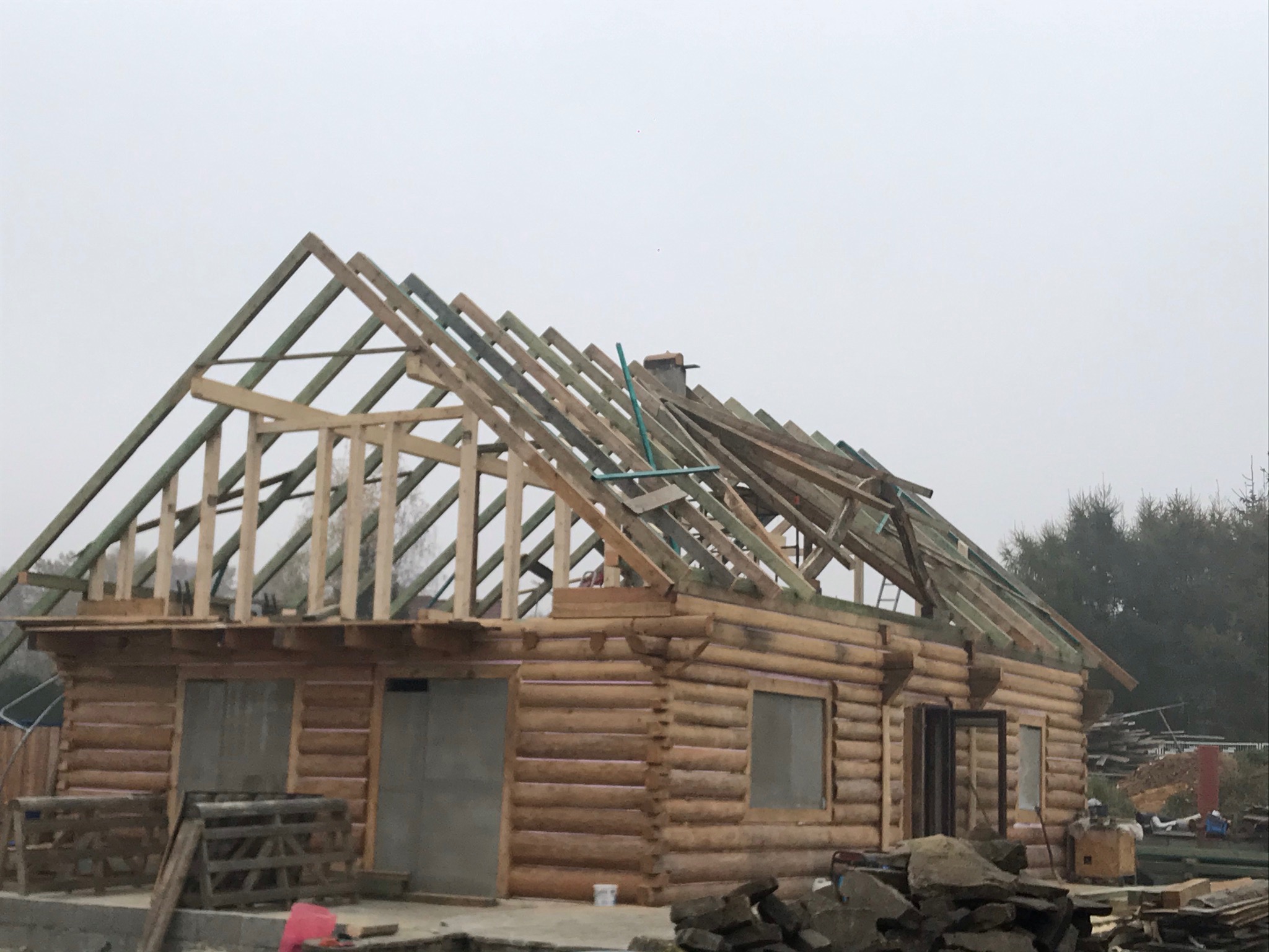 Budowa domu z bali w stanie surowym otwartym, widoczne ściany z bali, konstrukcja dachu z drewnianych krokwi, w tle las i pochmurne niebo.
