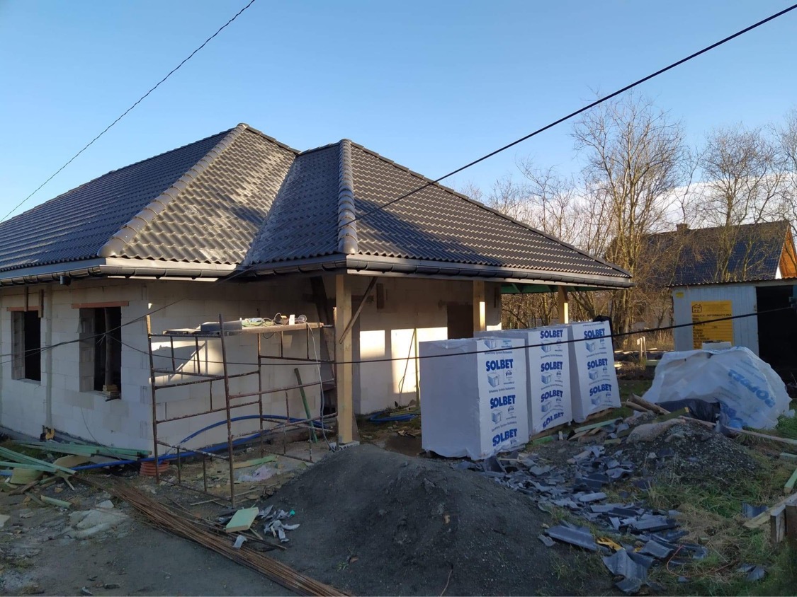 Budowa domu jednorodzinnego w trakcie realizacji, widoczny nowy, ciemny dach z dachówki, ściany z białego betonu komórkowego, rusztowanie, materiały budowlane Solbet i sterta ziemi.