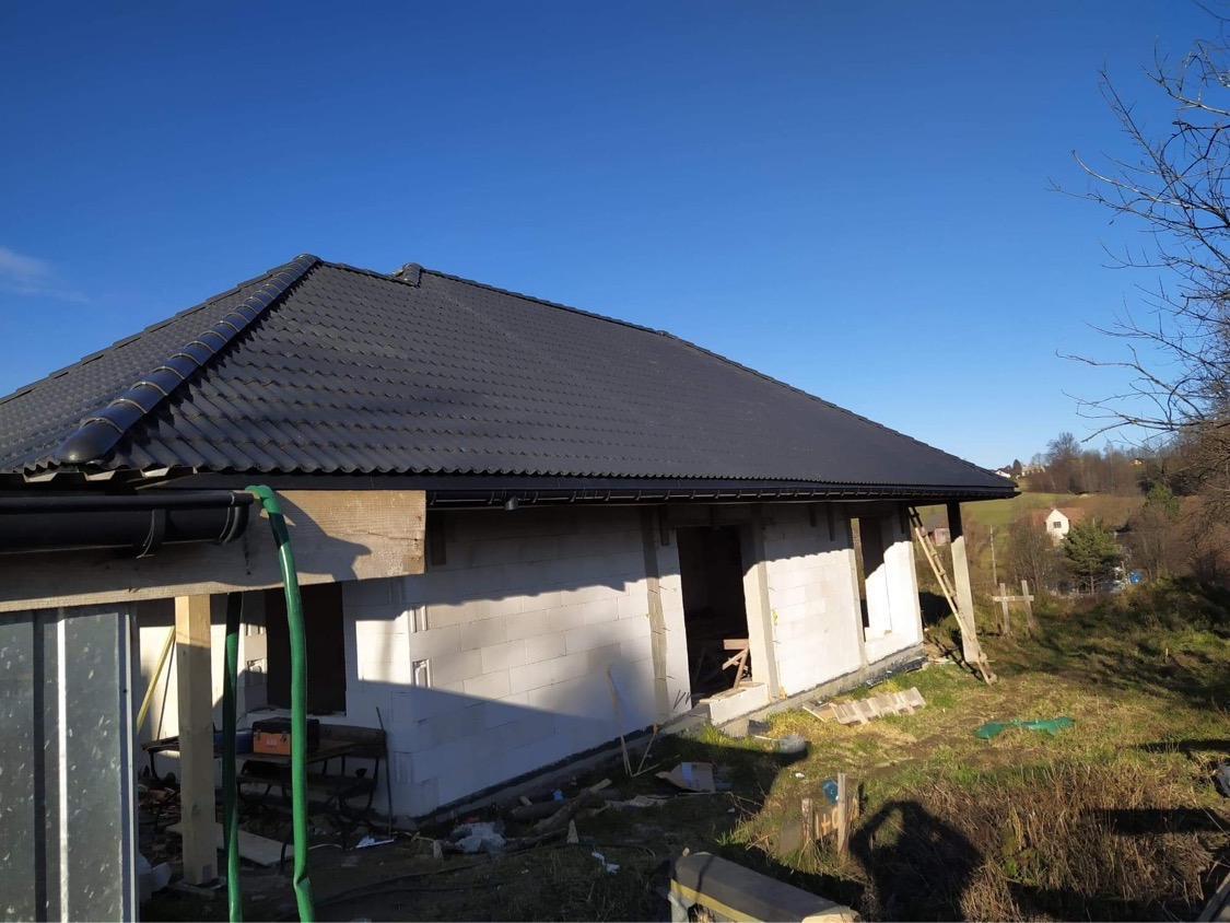 Niedokończony dom murowany z ciemnym dachem z blachodachówki, widoczne białe bloczki, rynna i fragment węża ogrodowego, w tle pagórkowaty krajobraz.