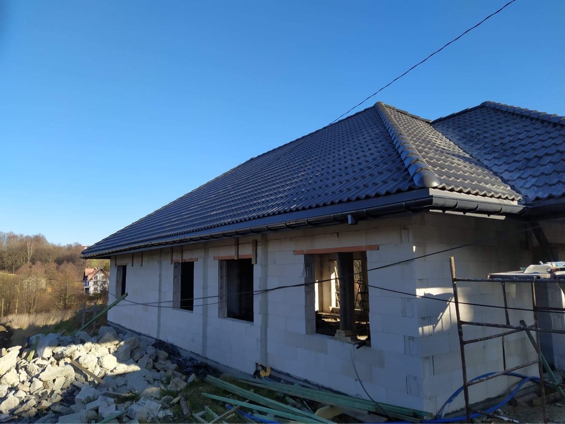 Dom w trakcie budowy: ściany z białych bloczków, ciemnoszary dach z rynną, widoczne otwory okienne, gruz i materiały budowlane na pierwszym planie, błękitne niebo w tle.