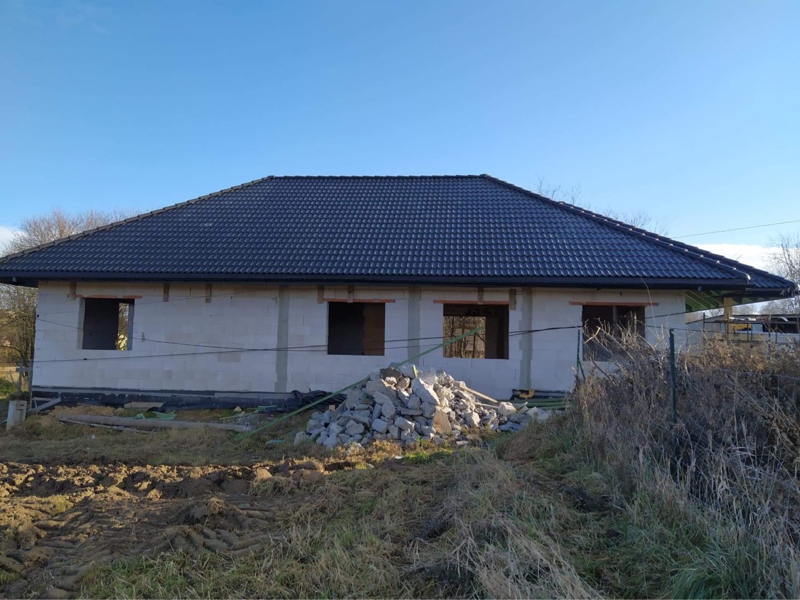 Surowy stan domu jednorodzinnego z ciemną dachówką, widoczne otwory okienne i stos gruzu przed budynkiem.