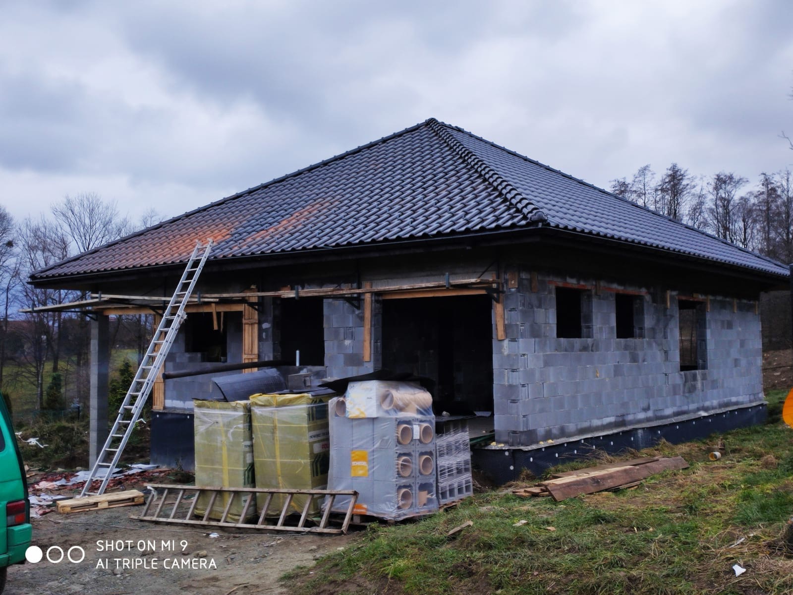 Niedokończony budynek jednorodzinny z szarej cegły z założonym czarnym dachem z dachówki ceramicznej, widoczne rusztowania, drabina oparta o dach, materiały izolacyjne i wentylacyjne na paletach...