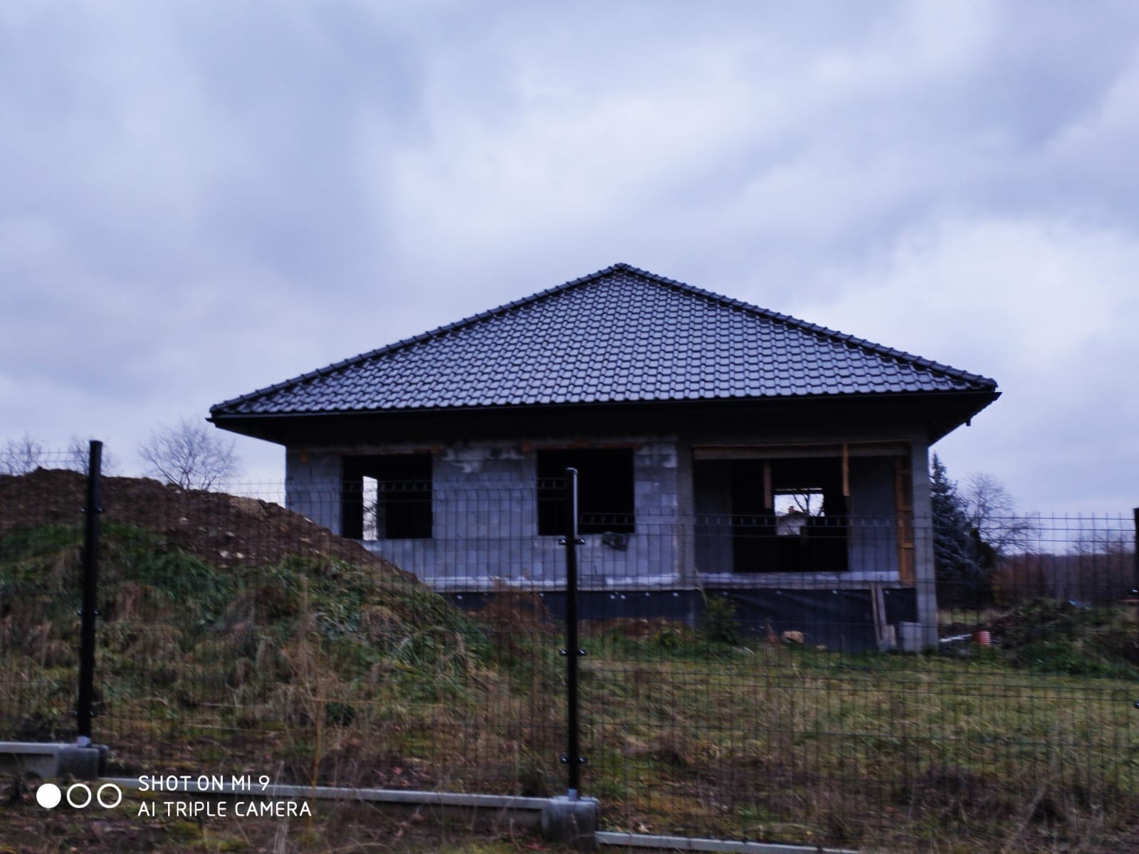 Surowy stan domu murowanego z ciemnym dachem, widoczny przez ogrodzenie z siatki na tle pochmurnego nieba. Budynek w trakcie budowy, bez okien i drzwi, z widocznymi bloczkami. Na pierwszym planie...