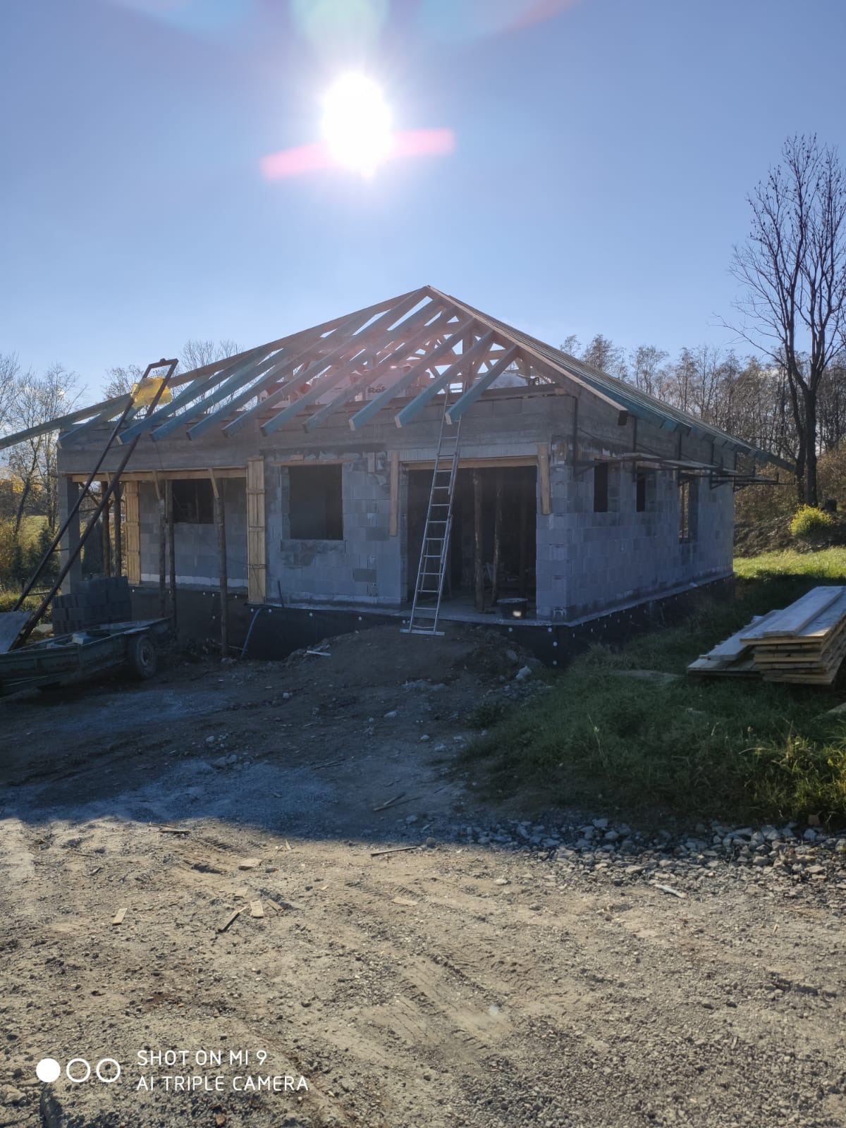 Wznoszony dom z szarego bloczka z widocznym drewnianym szkieletem dachu w słoneczny dzień, z drabiną opartą o ścianę i stertą desek obok.