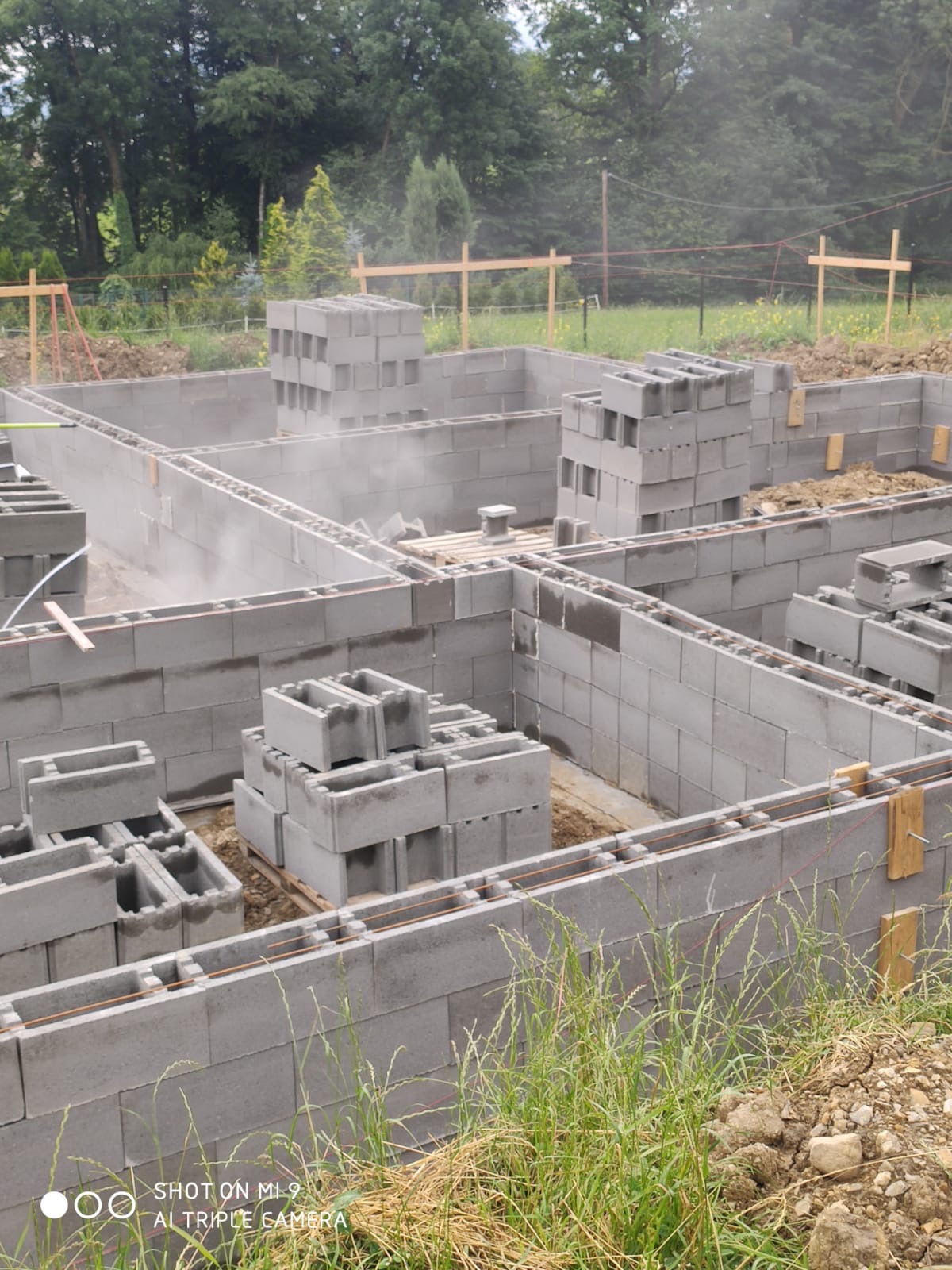Fundamenty domu w trakcie budowy, widać szare bloczki betonowe ułożone w rzędach, wystające pręty zbrojeniowe, drewniane elementy szalunku i sterty bloczków czekające na wmurowanie.